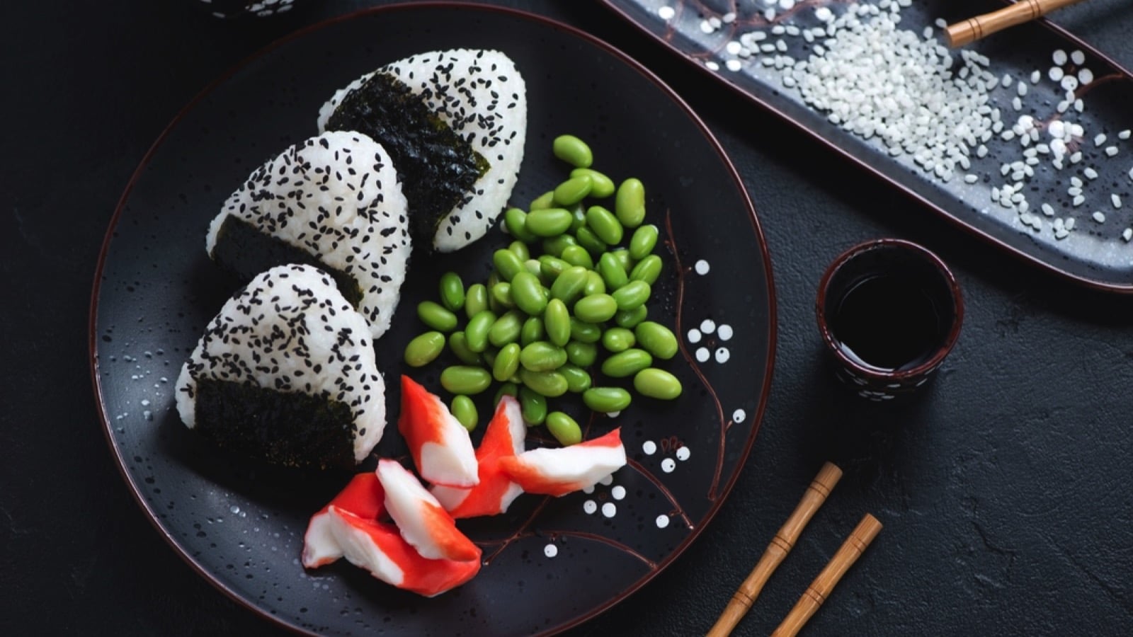 Plate with onigiri or japanese rice balls, crab meat and edamame, high angle view on a black stone background, horizontal shot